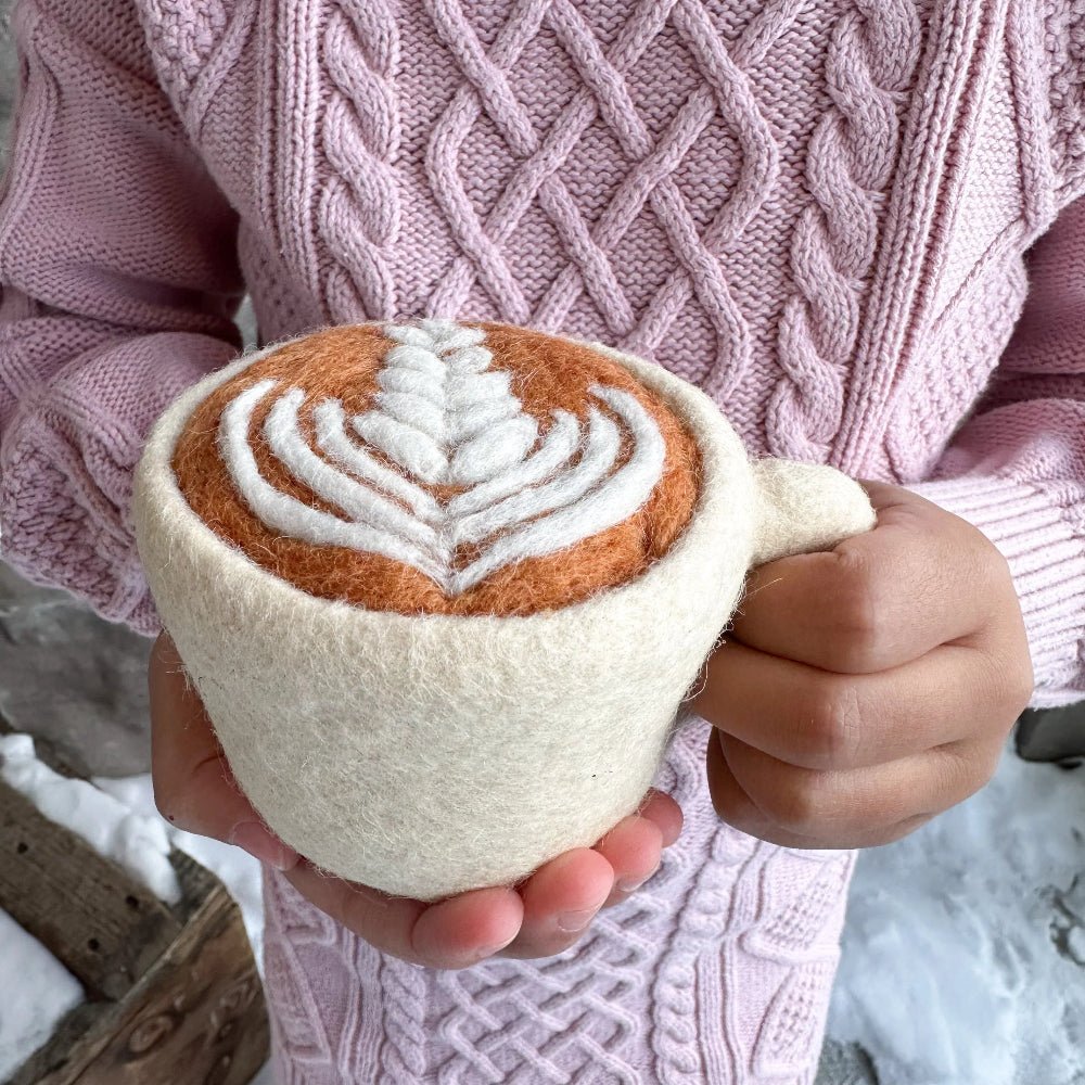A person in a pink cable-knit sweater holds a hand-felted mug shaped like a cappuccino. The mug has a cream-colored felt body and handle, with a felted coffee surface in warm brown and white leaf-latte pattern, set against a snowy outdoor background.