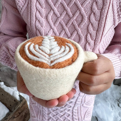 A person in a pink cable-knit sweater holds a hand-felted mug shaped like a cappuccino. The mug has a cream-colored felt body and handle, with a felted coffee surface in warm brown and white leaf-latte pattern, set against a snowy outdoor background.