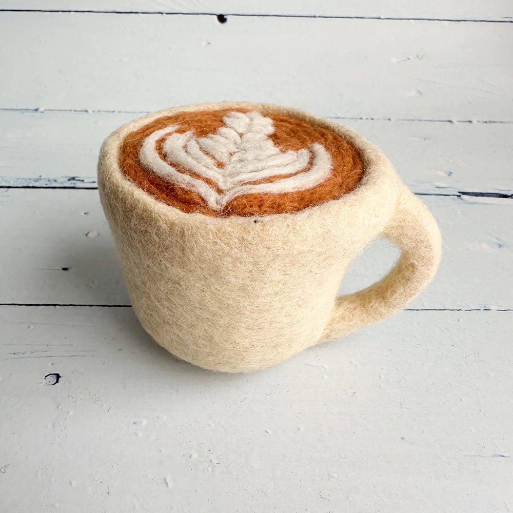 A hand-felted ceramic mug styled to look like a cappuccino, featuring a cream-colored felt body, a brown felted coffee surface, and a white felted latte art pattern, set on a white wooden plank surface.