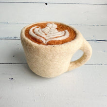 A hand-felted ceramic mug styled to look like a cappuccino, featuring a cream-colored felt body, a brown felted coffee surface, and a white felted latte art pattern, set on a white wooden plank surface.