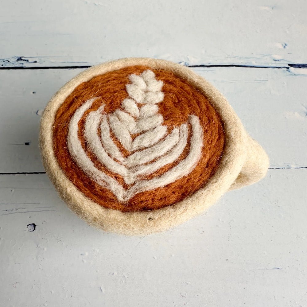 A handcrafted felted wool mug shaped like a cup of coffee with latte art, featuring a warm brown center and cream-colored leaf pattern, set against a light blue wooden background.