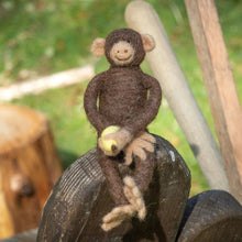 Needle felted monkey holding a banana perched on a wooden fence in a nature setting