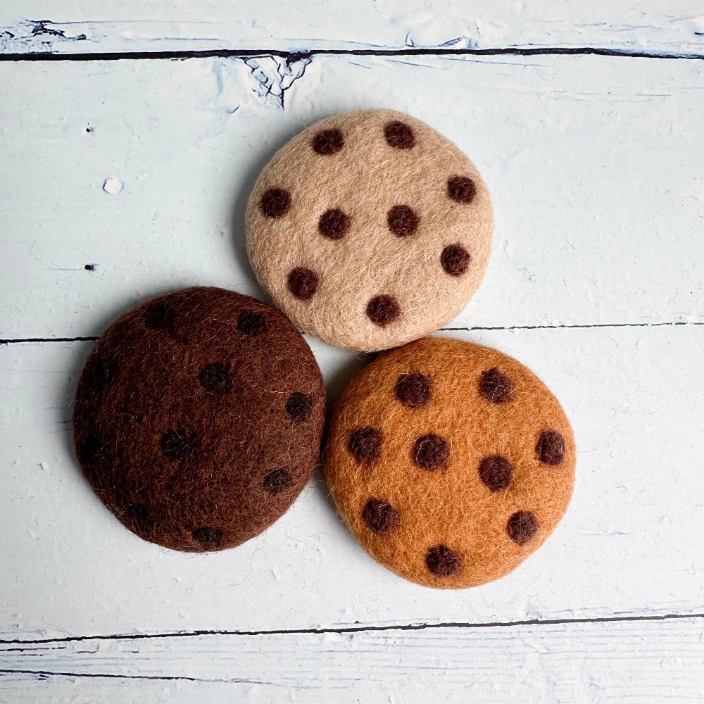 Three felt cookies in beige, brown, and orange-brown shades with dark chocolate chip details, arranged on a white distressed wood background.