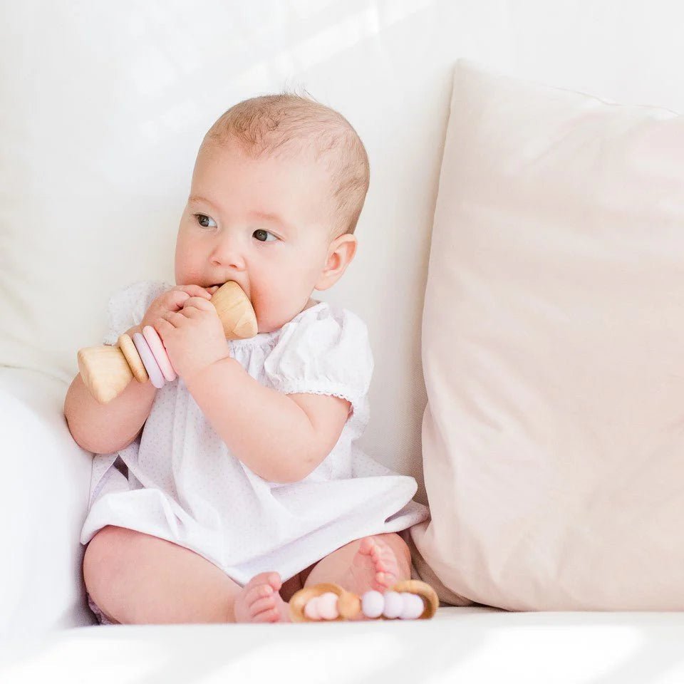 Baby in a white onesie holding a wooden baby rattle on a white couch.