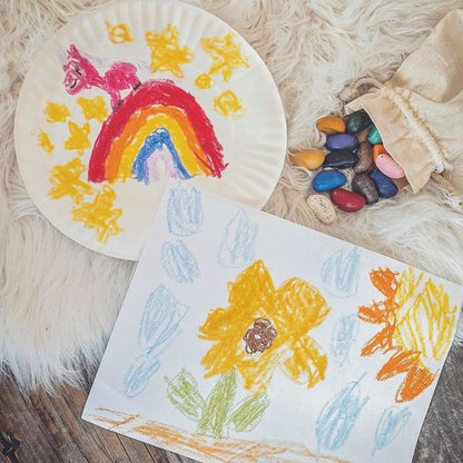 Children's drawings of a rainbow and flowers on paper, with colored rock-shaped crayons beside them.