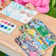 Children's watercolor paper with dinosaur designs on a wooden table, next to a watercolor set.