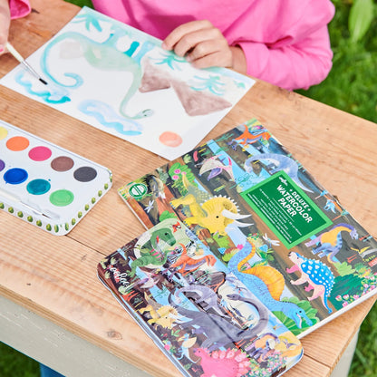 Children's watercolor paper with dinosaur designs on a wooden table, next to a watercolor set.