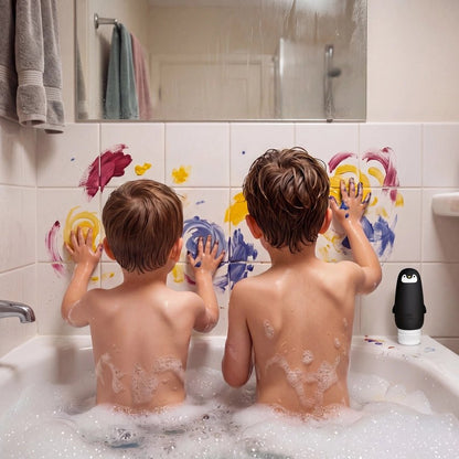 Two young boys sitting in a bubble-filled bathtub, seen from behind as they paint on a tiled bathroom wall with colorful paint splatters. A small black and white penguin toy sits on the tub's edge.