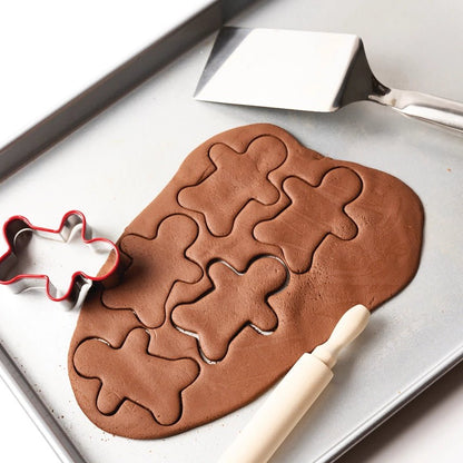 Gingerbread dough with gingerbread man shapes on a baking tray, with a spatula and cookie cutter.