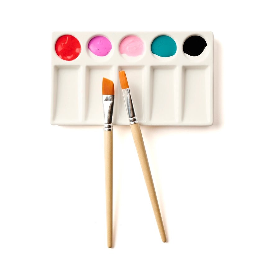 White ceramic paint palette with 5 wells filled with paint and two paint brushes lying on top on a white background