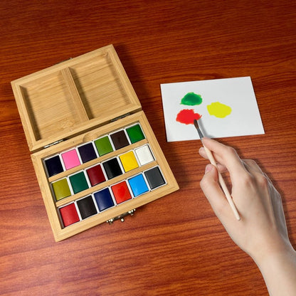 Hand dipping a paint brush into a palatte next to a watercolor set in a wooden box on a wooden surface