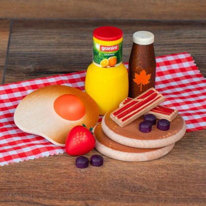 A wooden toy breakfast picnic set arranged on a red-and-white checkered cloth over a wooden surface. The set includes a wooden fried egg with a red yolk, a red strawberry, orange juice, maple syrup, two round wooden pancakes with striped bacon pieces, and five purple blueberries