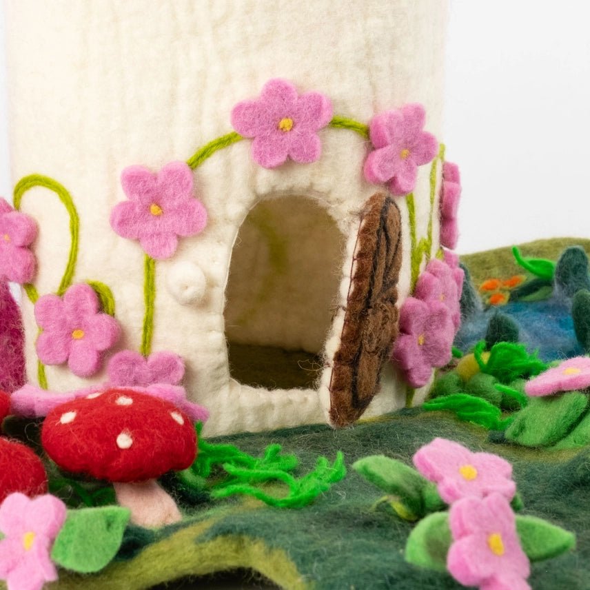 A close-up of a handmade felt forest house, featuring a cream-colored felt body decorated with pink felt flowers, green vines, a brown felt door, and colorful felt mushrooms and foliage.