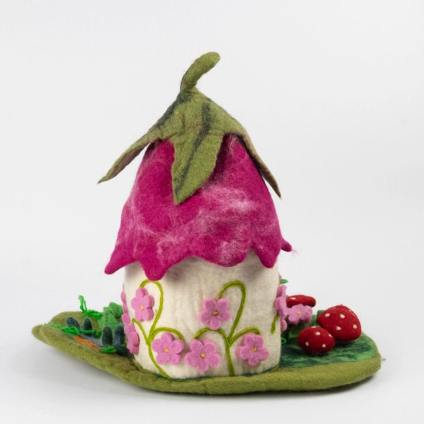The rear view of felted wool fairy house, featuring a bright pink felt roof with green leaf details, a cream-colored body decorated with pink flowers, and a green base with red strawberries, set against a plain white background.