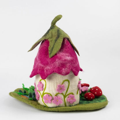 The rear view of felted wool fairy house, featuring a bright pink felt roof with green leaf details, a cream-colored body decorated with pink flowers, and a green base with red strawberries, set against a plain white background.