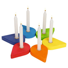 A set of six colorful wooden candle holders shaped like leaves in red, orange, yellow, green, blue, and dark blue, each holding a white taper candle with a flame, on a plain white background.