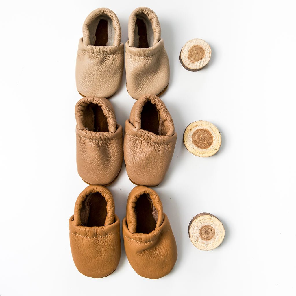 Three pairs of soft leather baby moccasins in beige, tan, and camel brown, arranged vertically next to three round wood slice props on a clean white background.