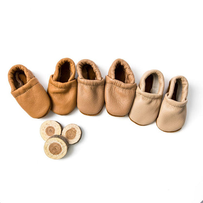 Six pairs of soft leather baby booties in various tan and beige shades, arranged in a row on a white background with three small round wood slice props at the bottom left.