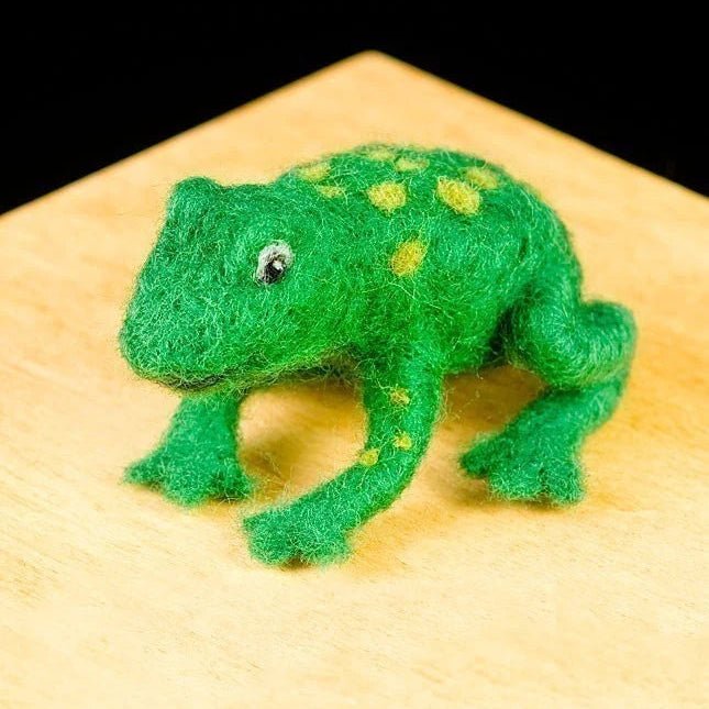 Green needle felted frog with light green spots on a wooden surface against a black background