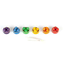 6 bowls in rainbow colors with matching wooden acorns lined up with wooden tongs in front