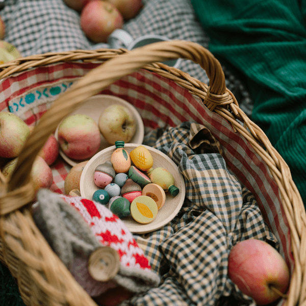 Wicker basket with apples, a checkered cloth, and colorful wooden fruits on a green surface.