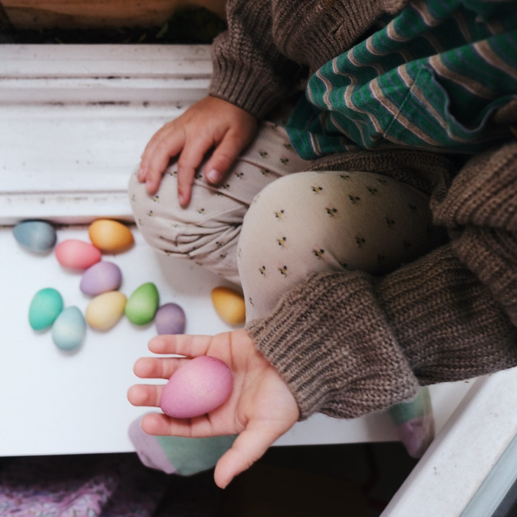Child holding a pink Easter egg with colorful eggs on a wooden surface