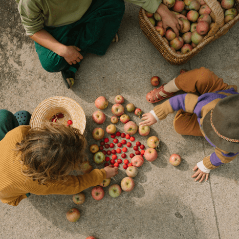 Three children sorting apples and red wooden half spheres on a concrete surface with a basket of apples nearby.