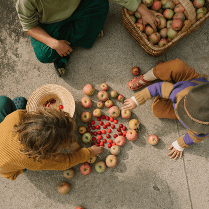 Three children sorting apples and red wooden half spheres on a concrete surface with a basket of apples nearby.