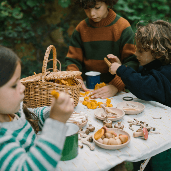 Children outdoors at a table with a basket, plates of food, real and wooden mushrooms