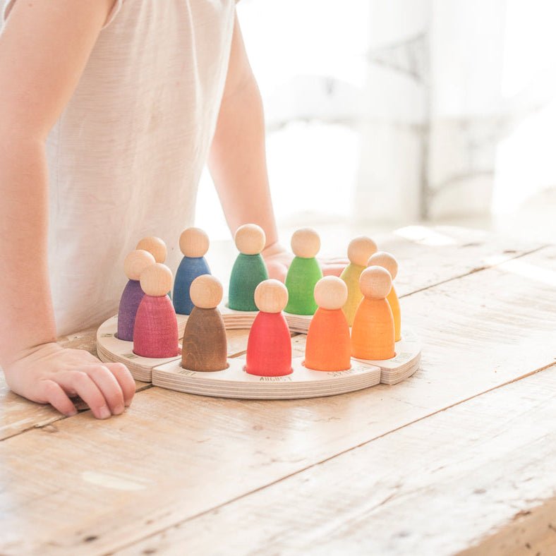 child standing over a wooden perpetual calendar with 12 differently-colored peg people on a wooden surface