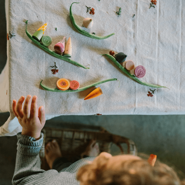 Children's hands reaching towards a table with wooden vegetables on a white cloth.