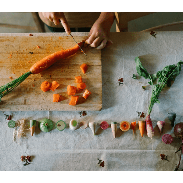 Child cutting a carrot on a wooden cutting board with various wooden vegetables arranged below.
