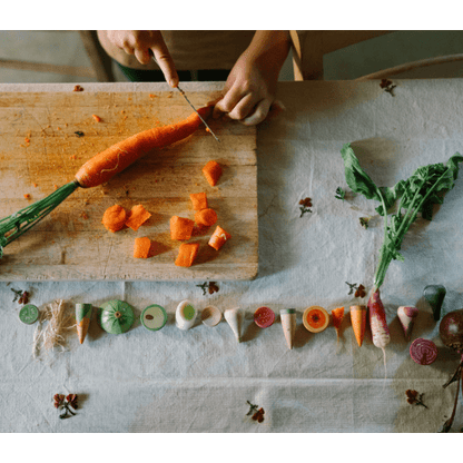 Child cutting a carrot on a wooden cutting board with various wooden vegetables arranged below.