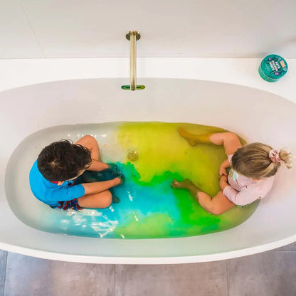 Two children in a bathtub filled with blue, yellow, and green water, with a tin of Honeysticks bath drops on the edge of the tub