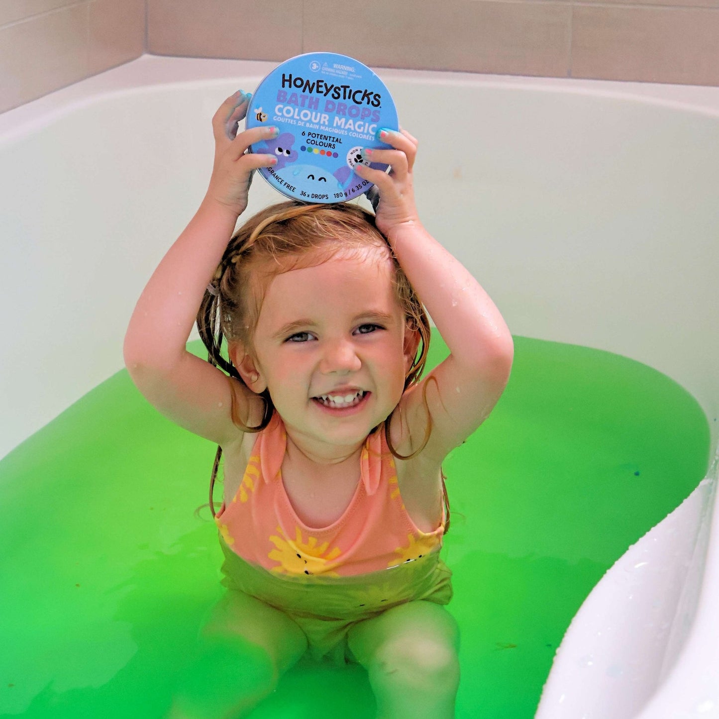 Child in a bathtub holding a tin of Honeysticks bath drops with green water.
