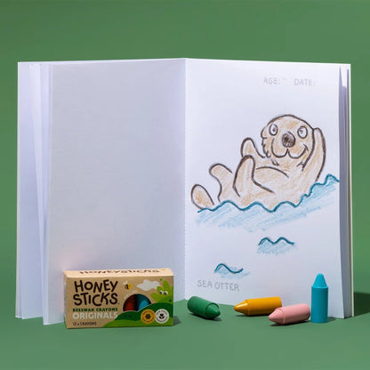 Honeysticks coloring book open to page of an otter, surrounded by Honeysticks crayons on a green background