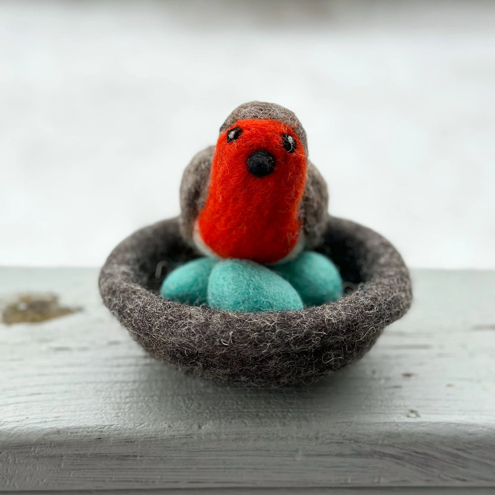 A small felted wool bird figurine with a bright red-orange chest, gray wings and head, and turquoise-blue eggs in a dark brown felt nest, placed on a light gray wooden surface with a soft blurred background.