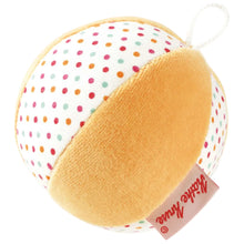 A round plush ball toy with a warm yellow-orange velvety side and a white fabric side covered in small multicolored polka dots. A small fabric tag with the brand name 'Kathe Kruse' is attached near the bottom.
