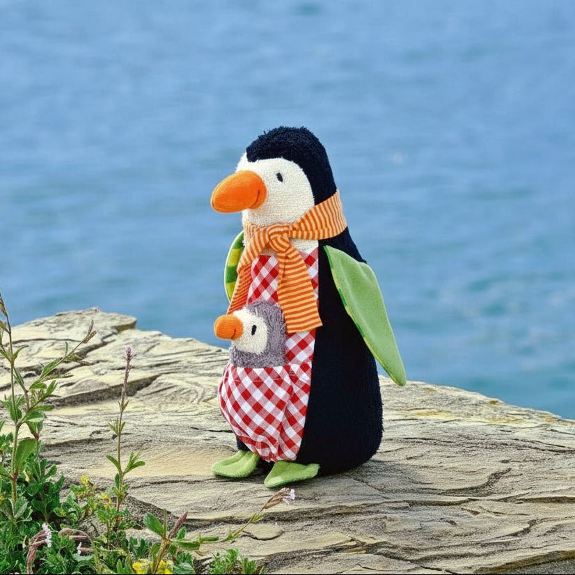 Plush penguin toy and gray baby with colorful patterned clothing and scarves are posed on a stone ledge outdoors with blue water and greenery in the background