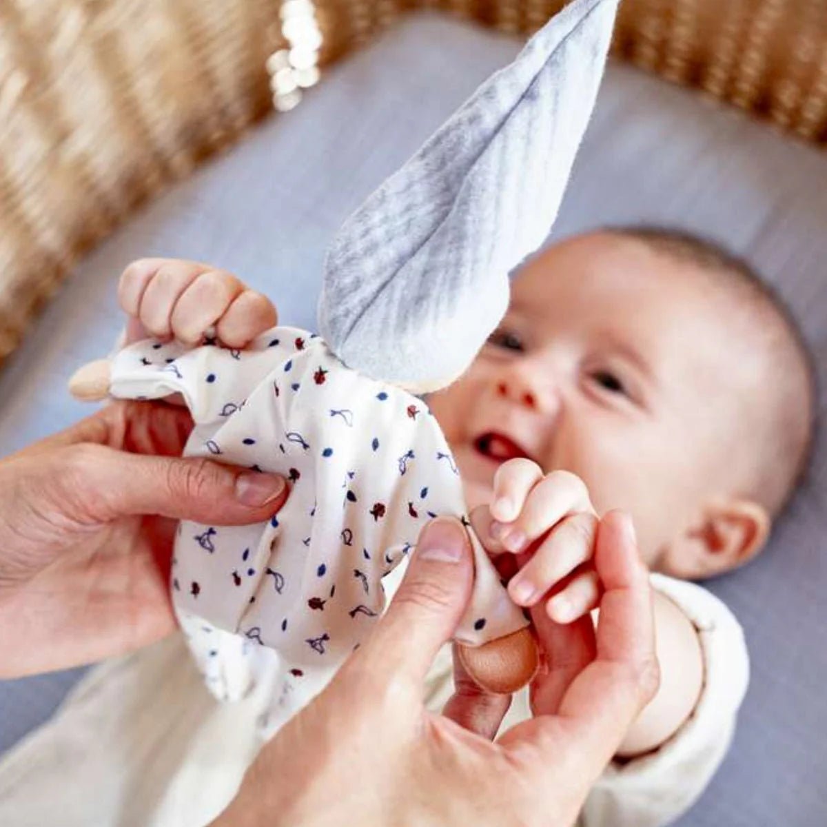 Baby lying on a crib with a person holding a small cloth doll wearing white pajamas with birds and ladybugs