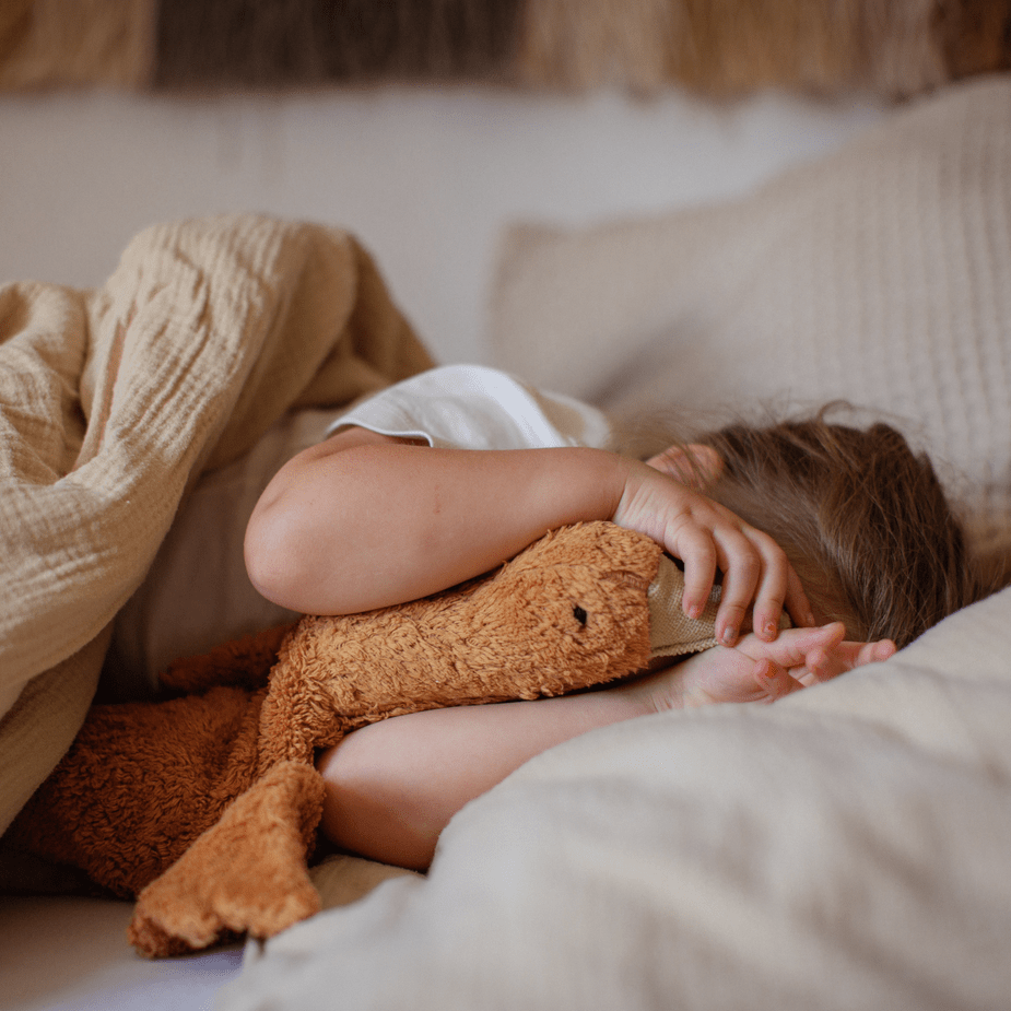 Child lying in bed under a blanket with a rust-colored stuffed goose, surrounded by soft pillows.