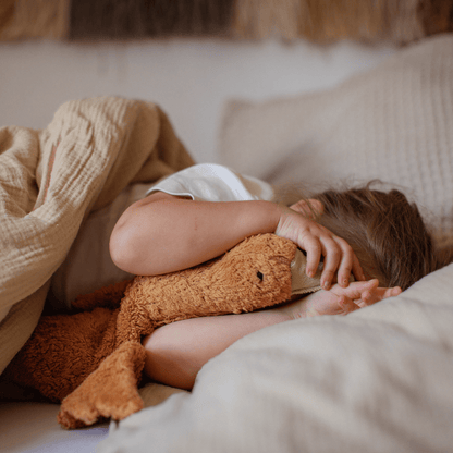 Child lying in bed under a blanket with a rust-colored stuffed goose, surrounded by soft pillows.