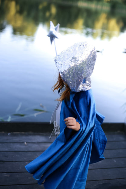 Child in profile on a wooden dock wearing a blue velvet cape and silver sequined hood, holding up a silver star wand