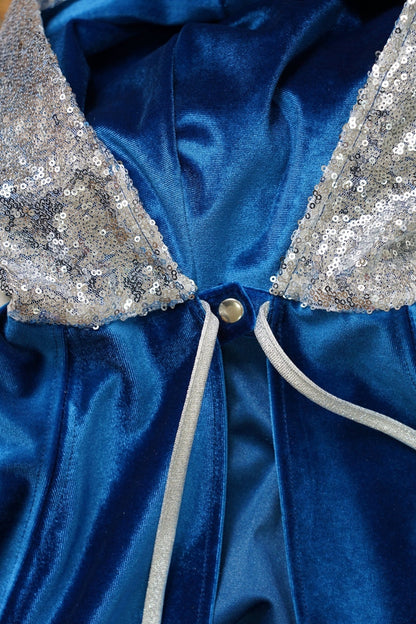 Close-up of the blue velvet cape neckline showing the silver sequined hood panels, metal snap, and silver ribbon tie