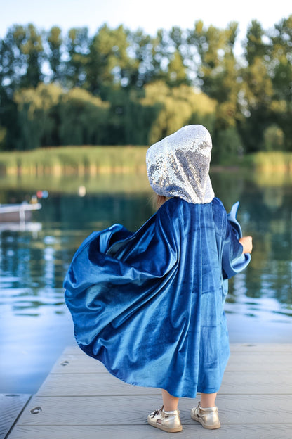 Child seen from behind on a lake dock wearing a flowing deep blue velvet cape with silver sequined hood