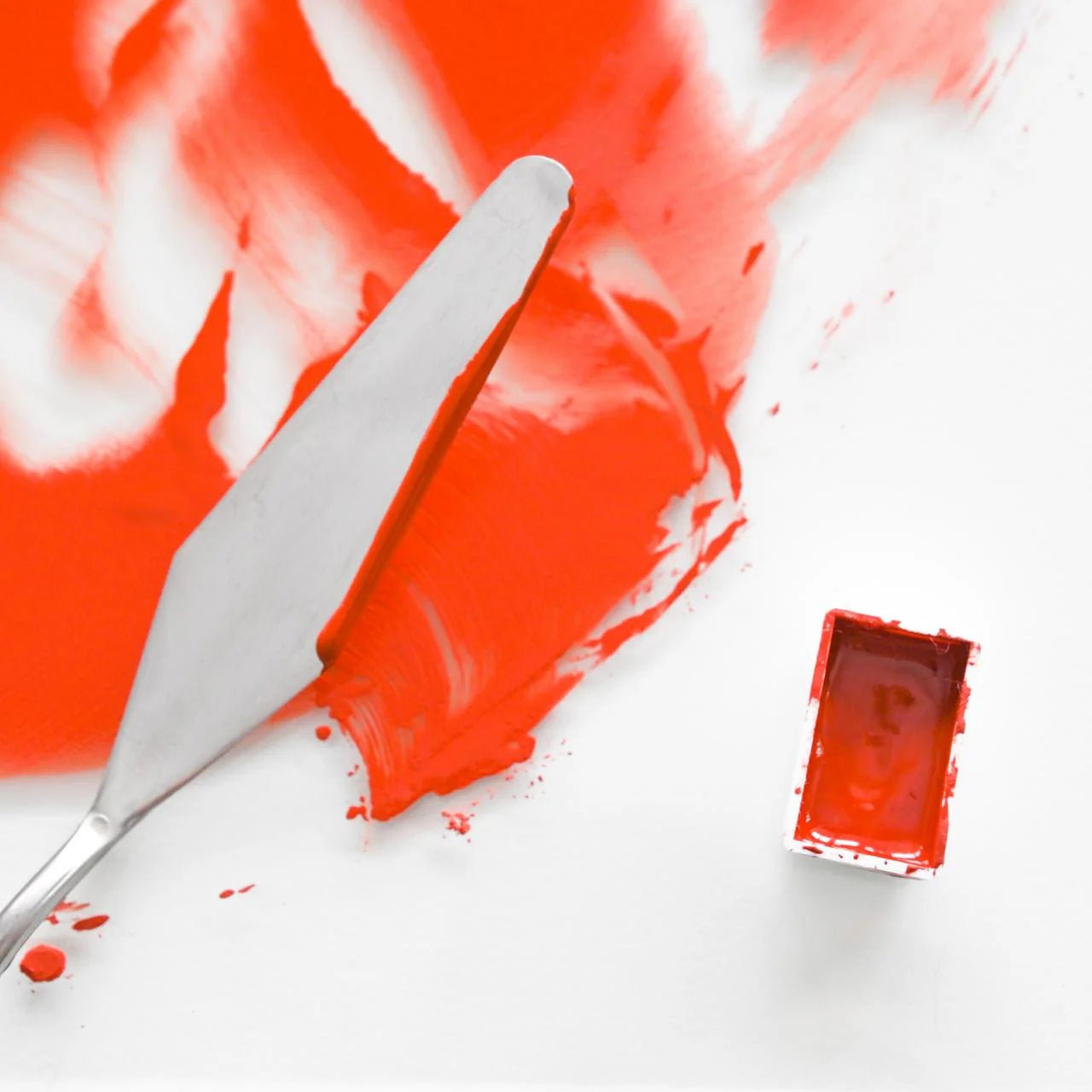Swatch of red watercolor paint on a white background with palette knife and palette.
