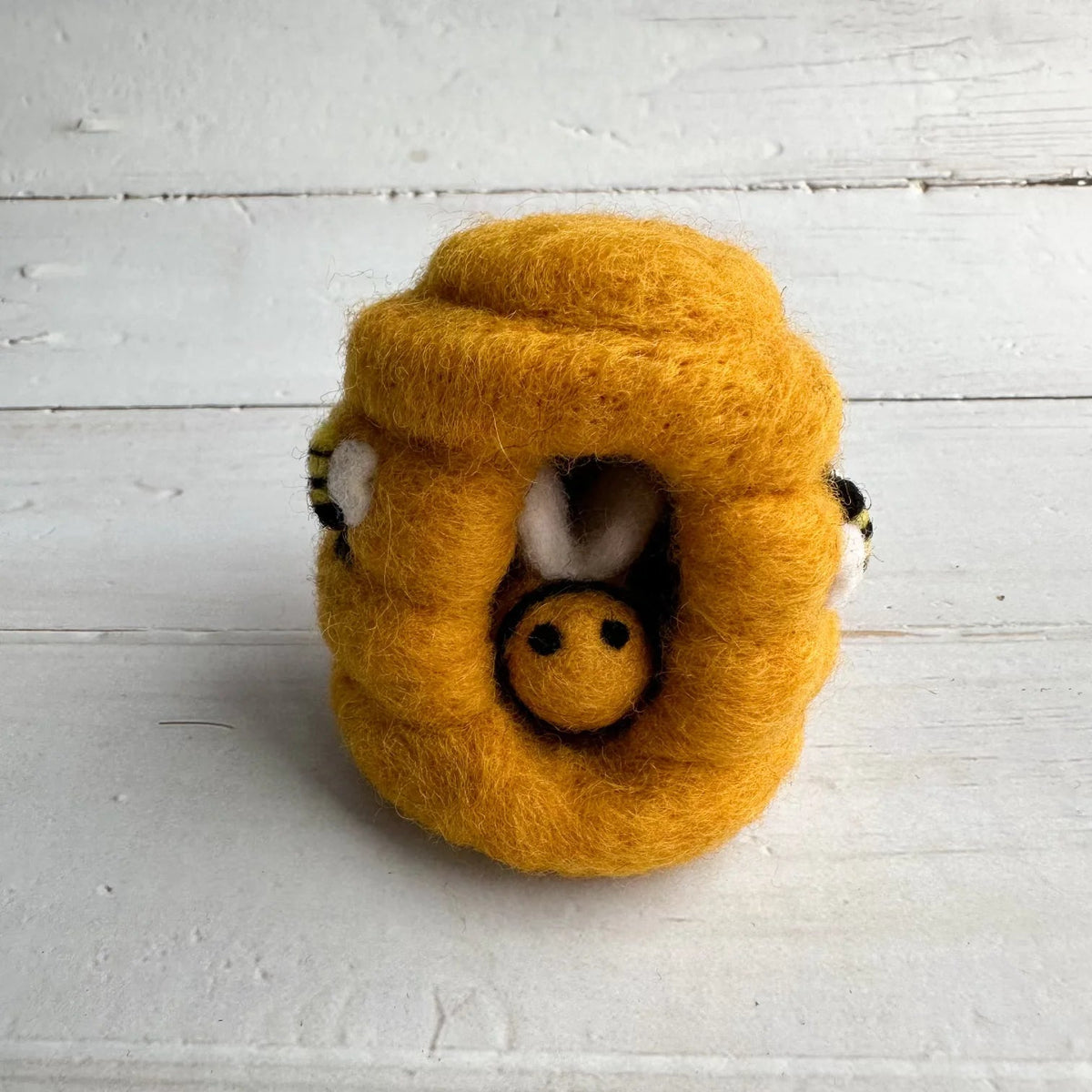 A yellow felted wool beehive toy with a small bee nestled inside, photographed on a white wooden plank surface.