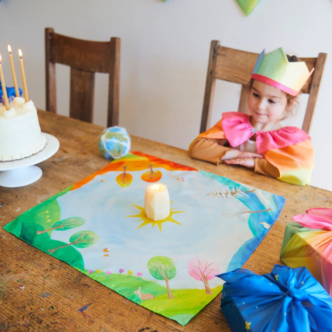 Child in a rainbow dress sitting at a table with a decorated cake and colorful seasonal playsilk with candle in the middle