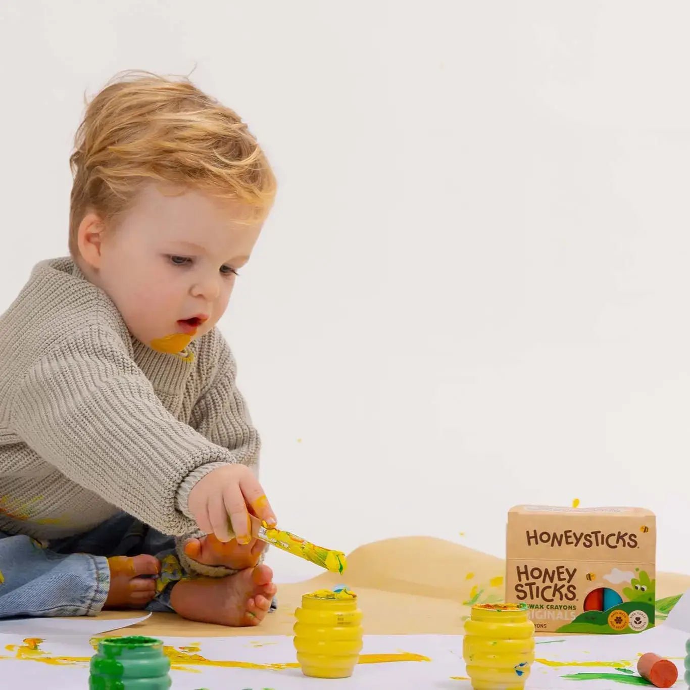Child playing with colorful Honeysticks paint on a white surface, with 'Honeysticks' packaging visible.