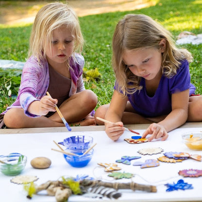 Two children sitting outdoors at a table with art supplies, including paint and brushes.